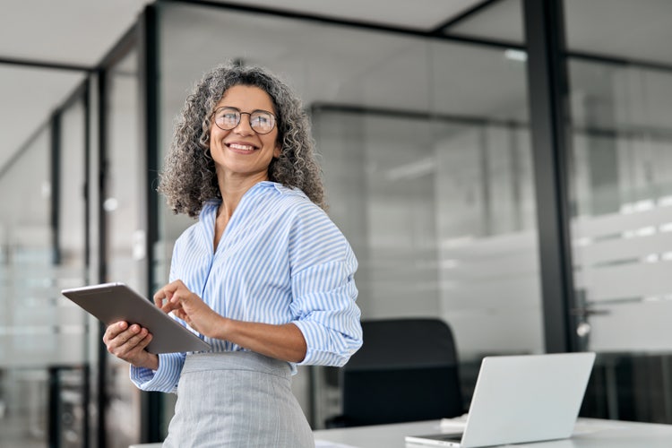 Smiling mature professional business woman bank manager, older happy female executive or lady entrepreneur holding digital tablet pad standing in office at work, looking away at copy space.